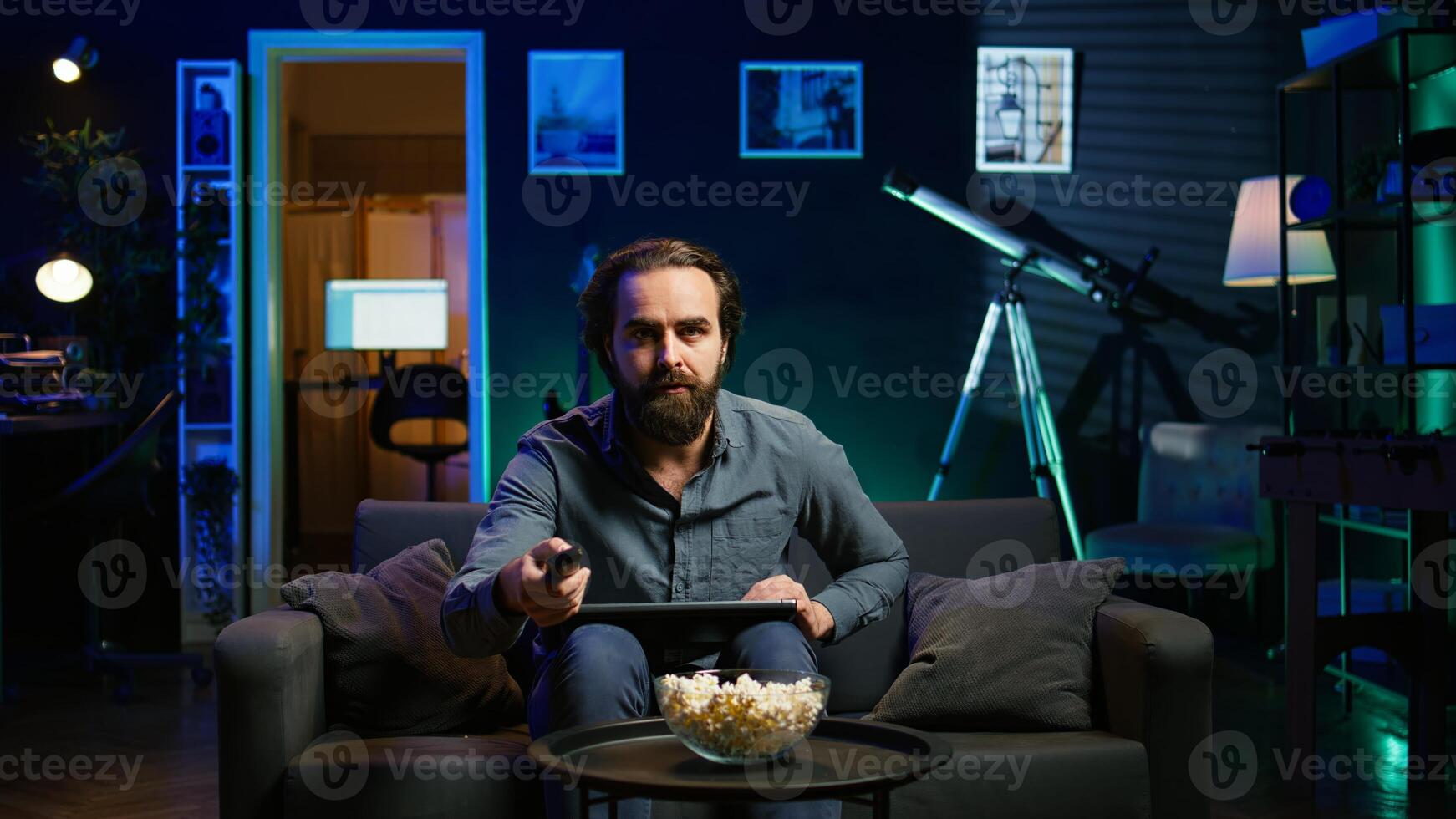 Man using laptop and watching breaking news on TV in neon lit apartment. Person turning volume on television, surprised by events while sitting on couch using notebook, camera B photo