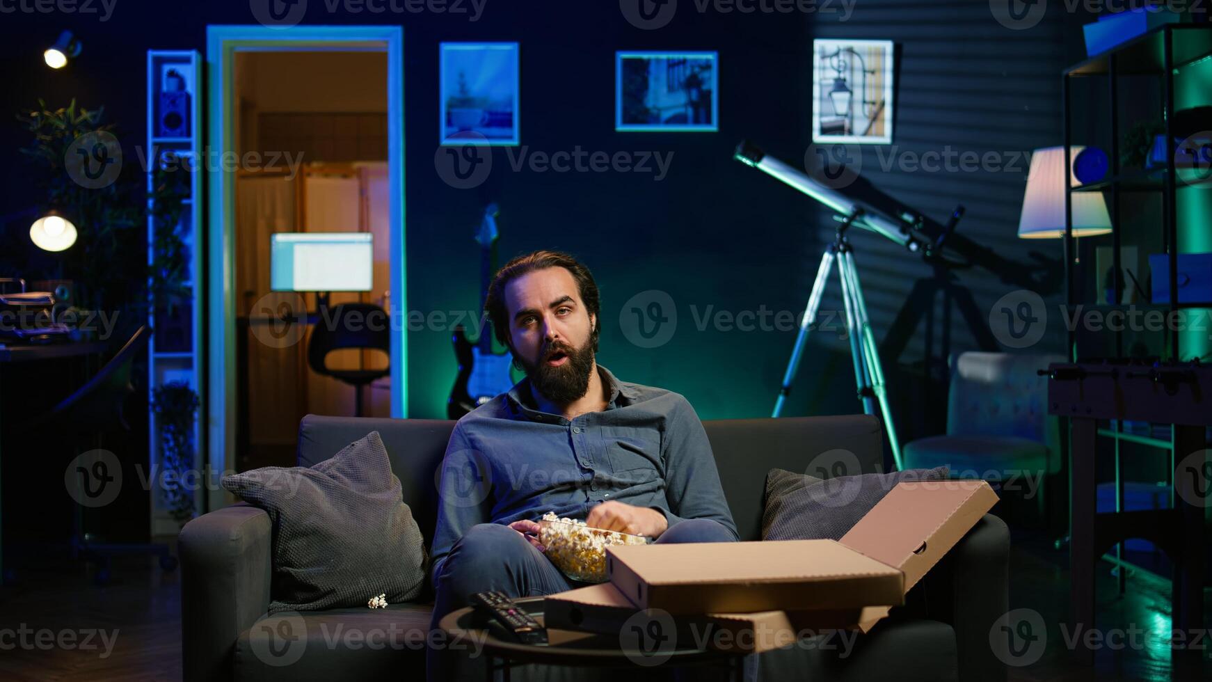 Exhausted man eating popcorn and watching TV late at night before going to sleep. Sleepy guy in pajamas eating snack on couch, consuming mindless entertainment, camera B photo