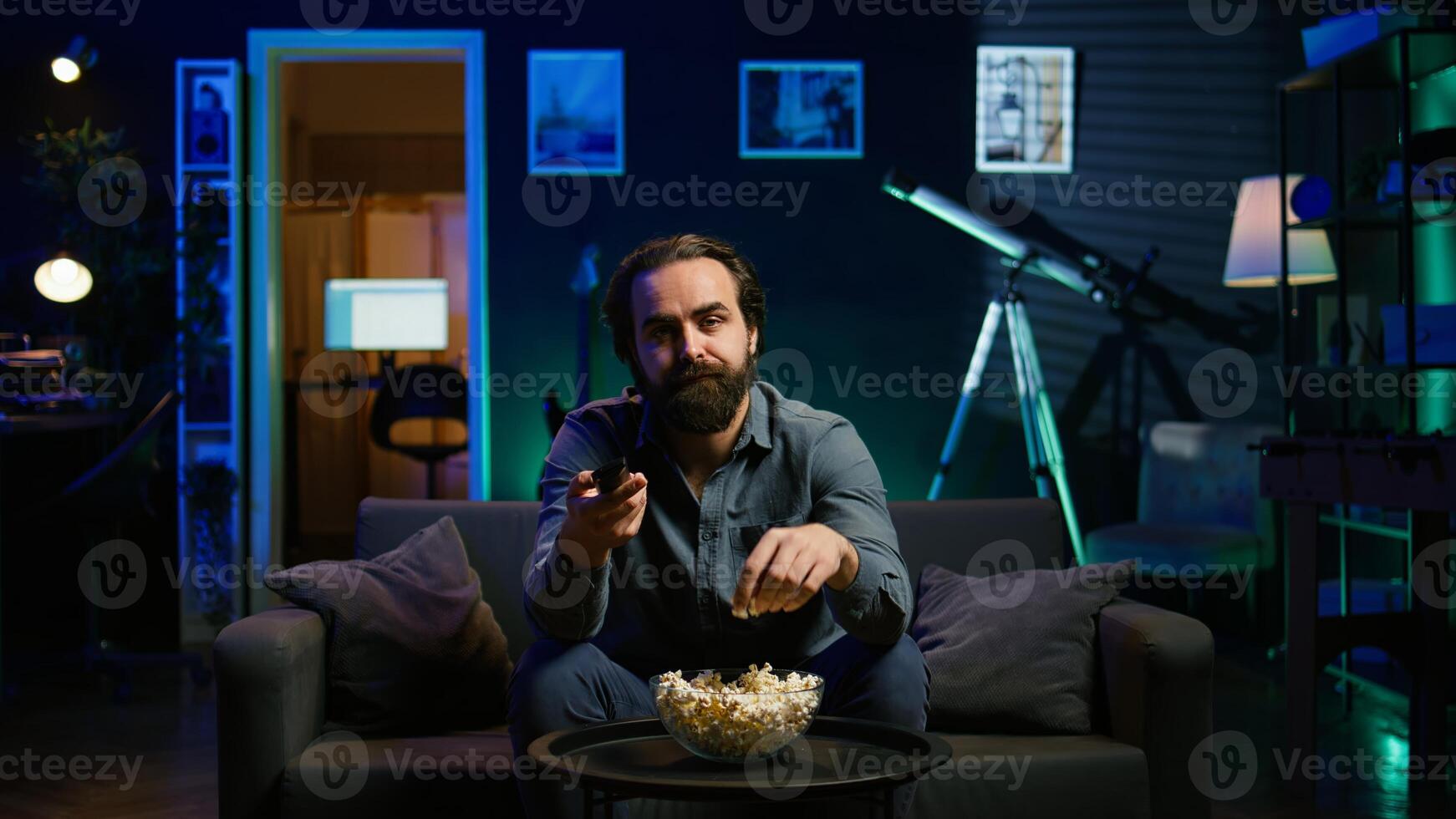 Bored man relaxing on couch, munching on popcorn while passing time watching movies on TV. Lazy person eating snacks and zapping through TV channels in RGB lit living room, camera B photo