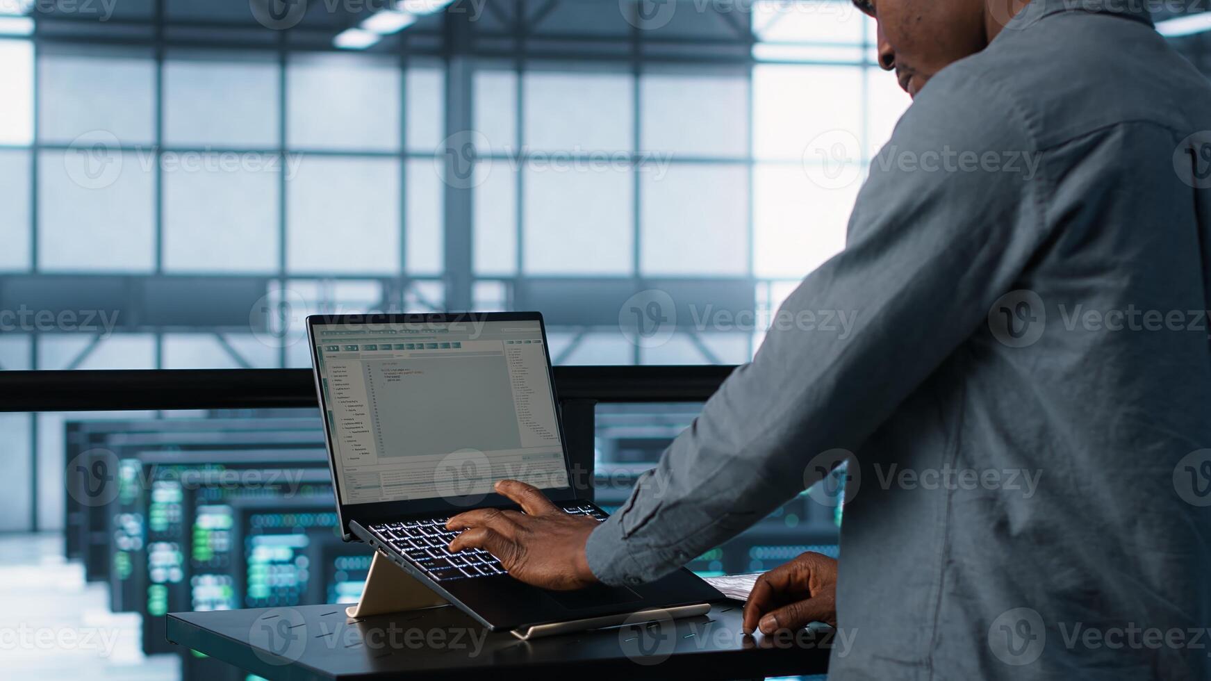 Computer scientist coding on laptop, developing disaster recovery plans. African american man in data center testing failover scenarios using notebook, camera A photo