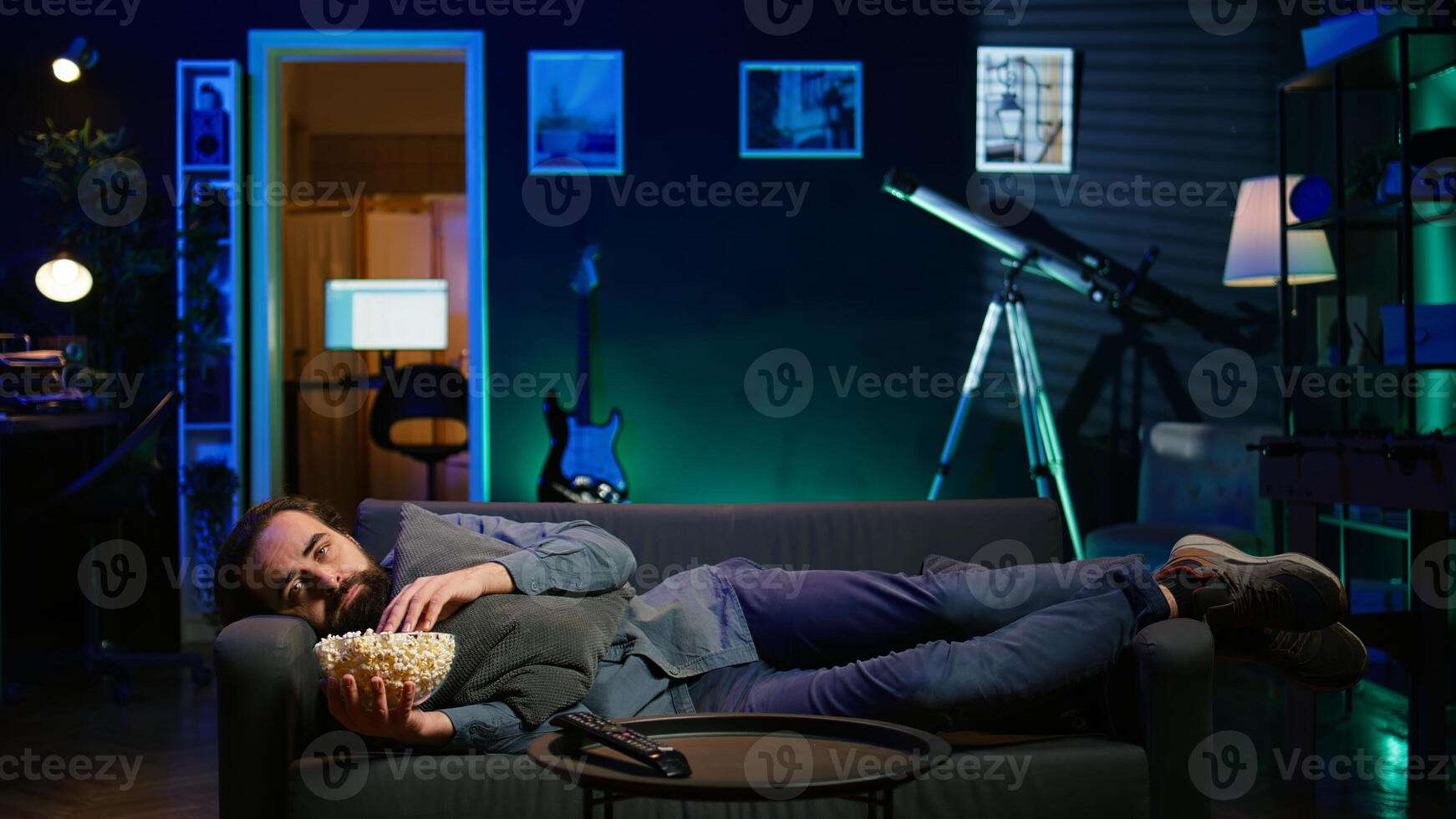 Depressed man at home lounged on couch feeling low, seeking comfort by watching movies and eating popcorn. Glum person laying down, suffering from depression, watching TV to feel better, camera B photo