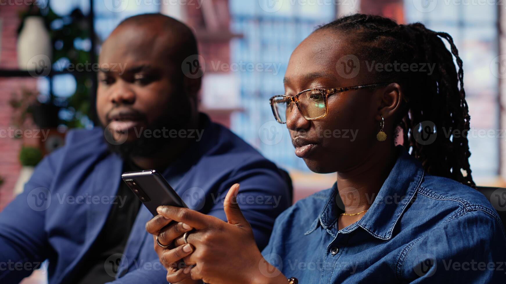 Black couple sitting together at home office and managing affairs in cozy loft. Young man solving work tasks on laptop and partner browses on smartphone. Camera B. photo