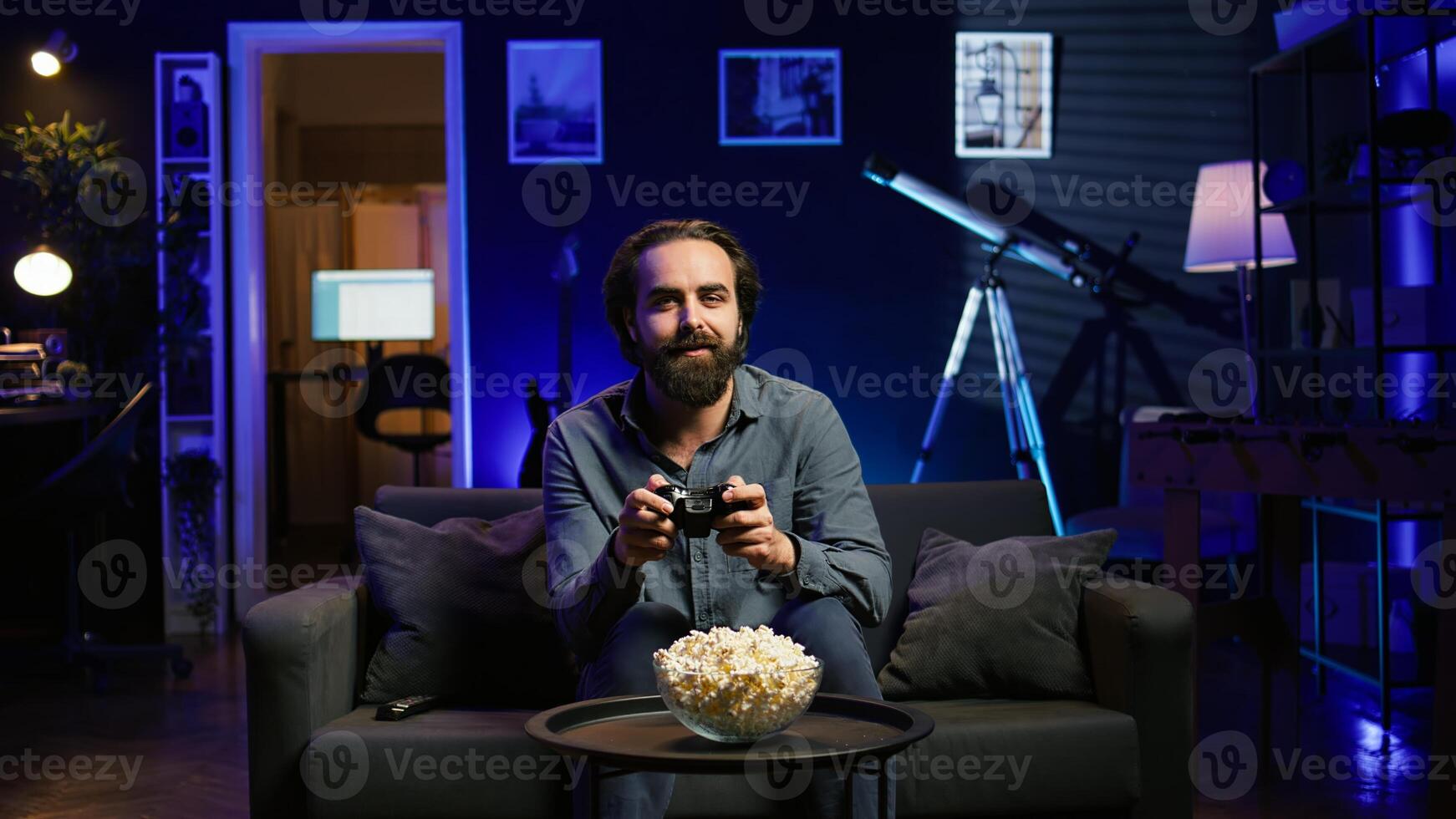 Excited man holds controller in neon illuminated apartment, playing videogames. Happy gamer on couch using joystick, participating in PvP game on console system, relaxing and having fun, camera B photo