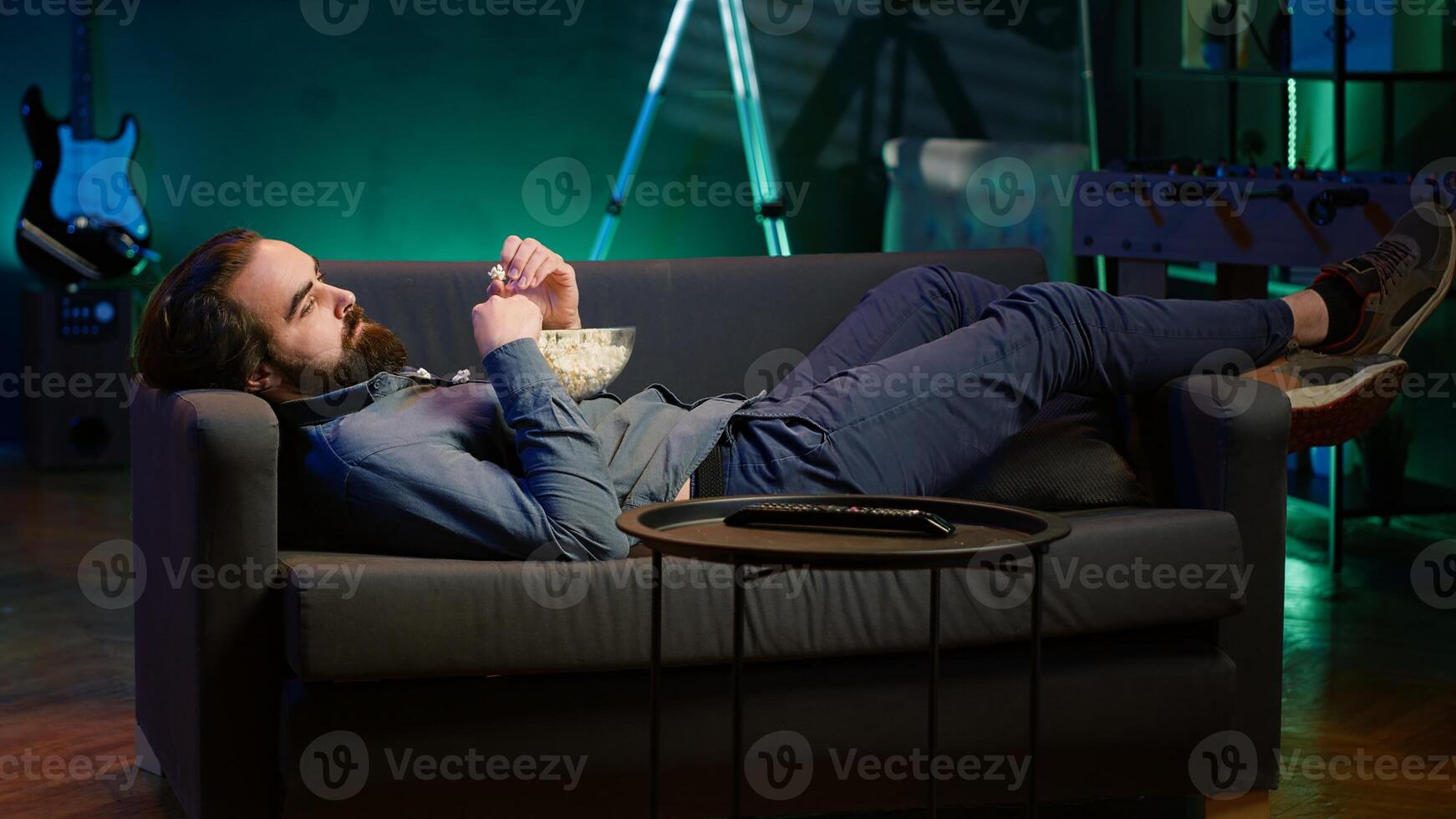 Man procrastinating at home, laying down on couch, munching on snacks and watching TV. Bored person living sedentary lifestyle, lounging on couch, wasting time watching TV, eating popcorn, camera A photo