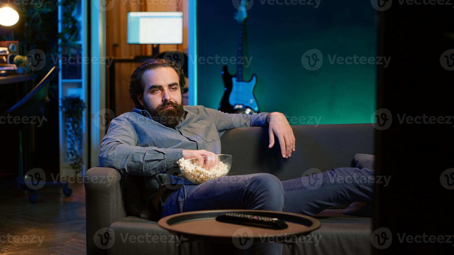Homeowner bored at home munching on snacks, using TV to binge series on streaming services. Man sitting on couch, wasting time with VOD shows on TV, eating popcorn, camera A photo