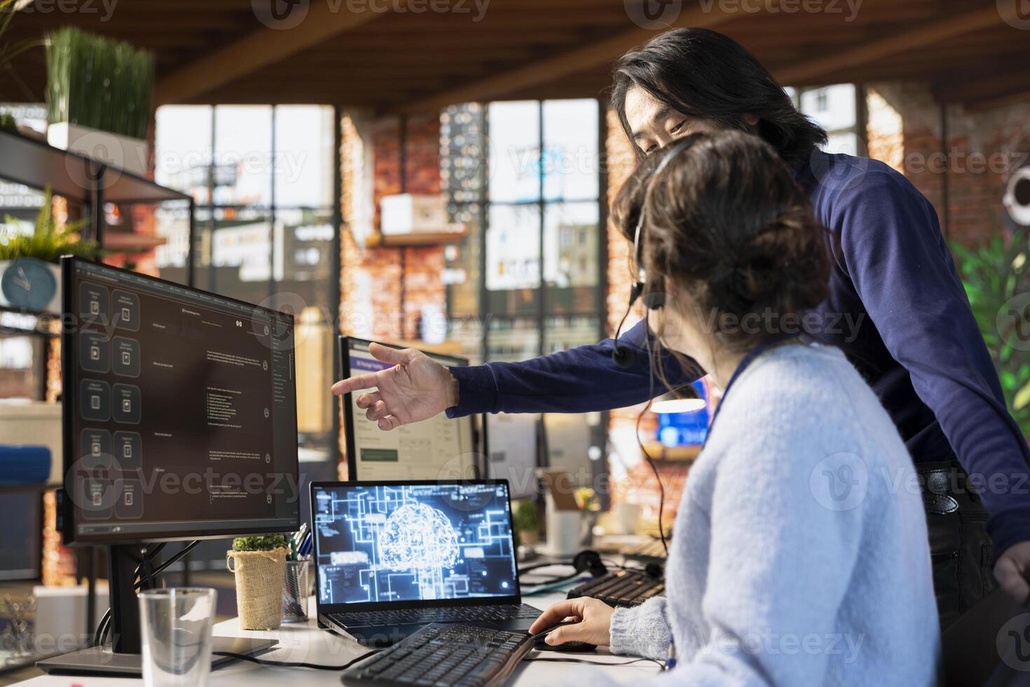 Call center manager guiding intern on how to use AI chatbot at office desk on PC. Asian customer support workplace onboarding specialist teaching woman to use artificial intelligence software photo