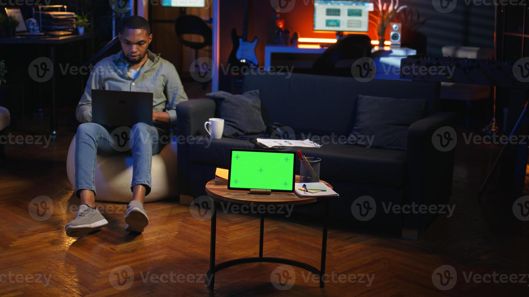 African american male freelancer enjoys free time next to chroma key on the bean bag, uses device to watch a podcast with headphones. Young guy relaxing remotely in the living room. Camera A. photo