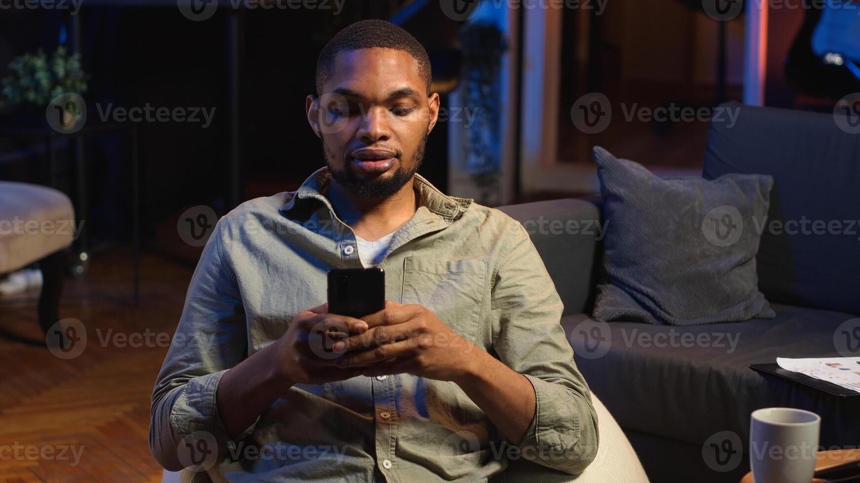 Young black guy texting his friends over social media messaging apps, using his smartphone to scroll on the internet. Focused person relaxing with his phone, sitting in his living room. Camera B. photo
