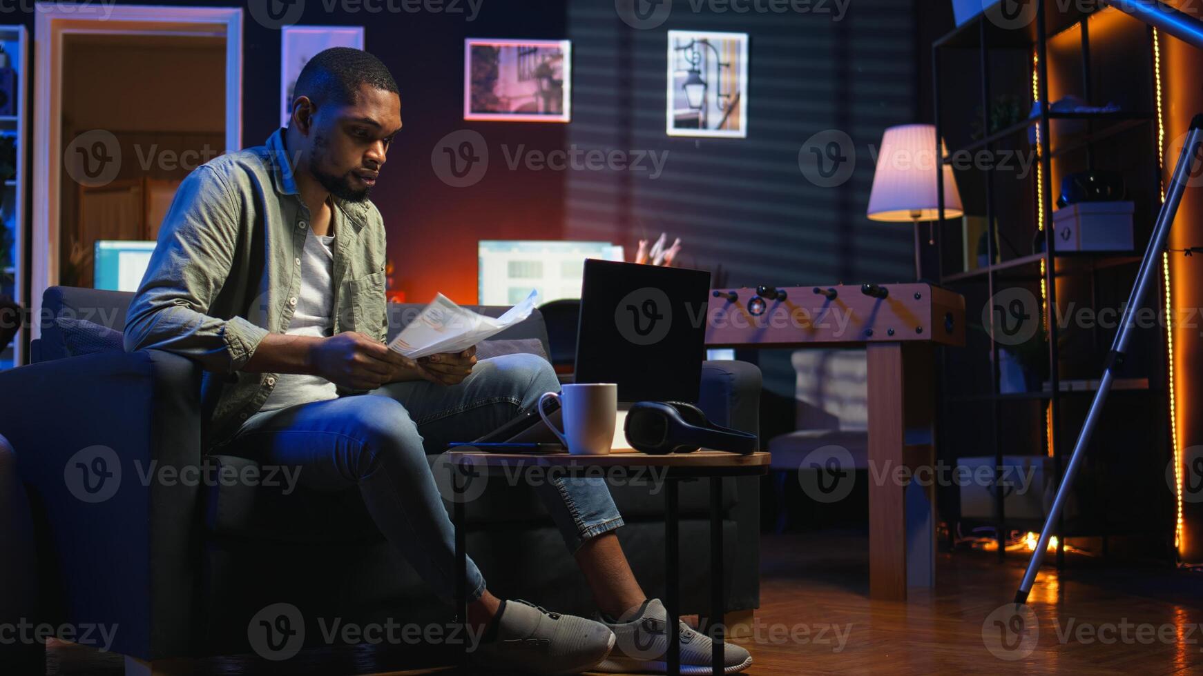 Black freelancer sitting on sofa and reading some papers to do research, multitasking with archives in the living room. Adult diving into professional or personal topics of improvement. Camera A. photo