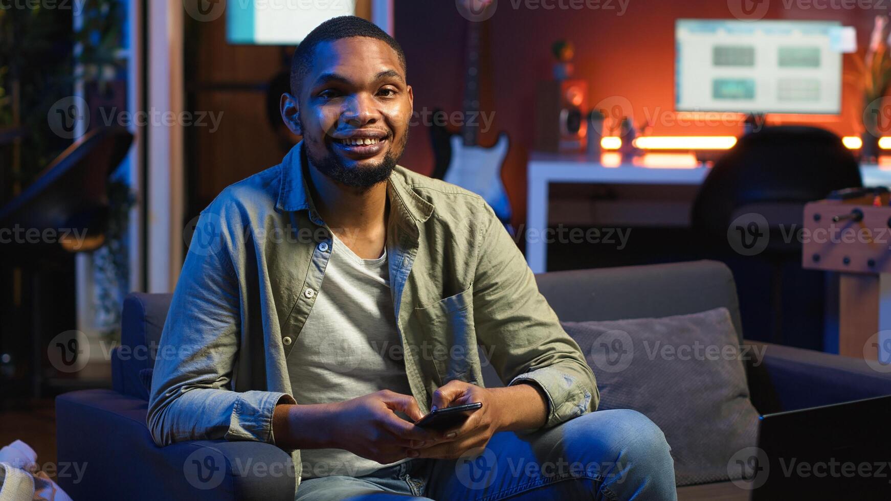 Smiling young adult chatting with friends over texts on mobile phone, sitting on couch and scrolling social media apps. Young guy staying entertained while working remotely. Camera B. photo