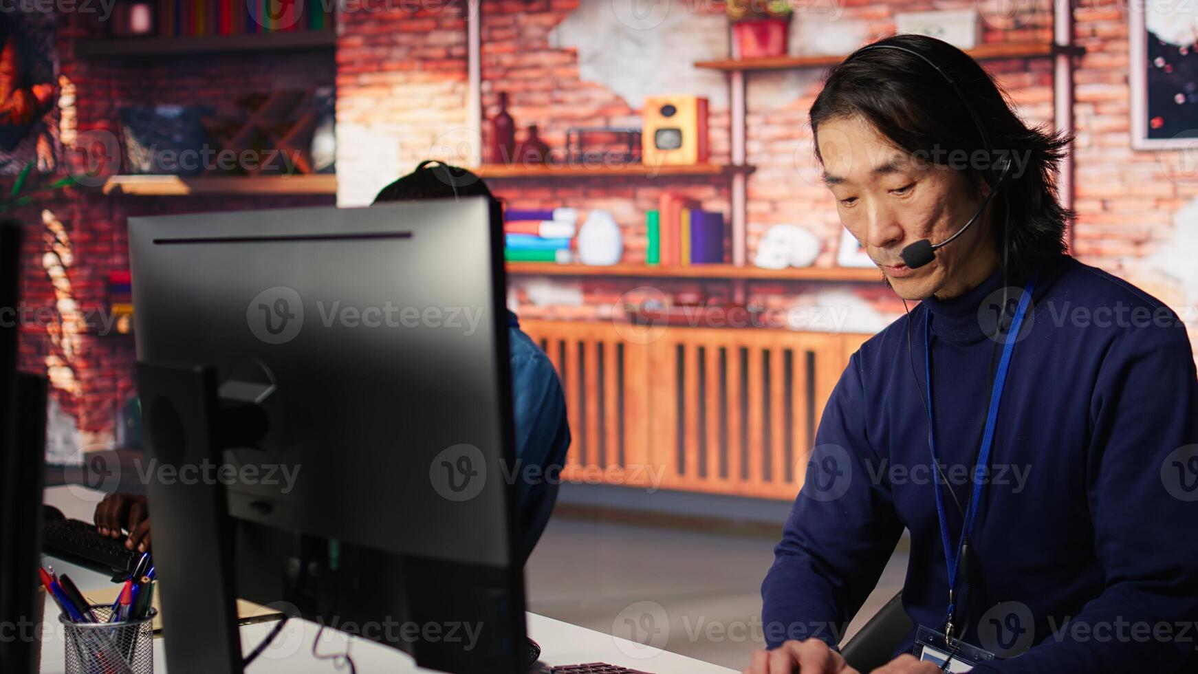 Call center coworkers dealing with upset customers, speaking with them online using headset at office desk. Asian man using PC, handling clients complaints, camera B photo