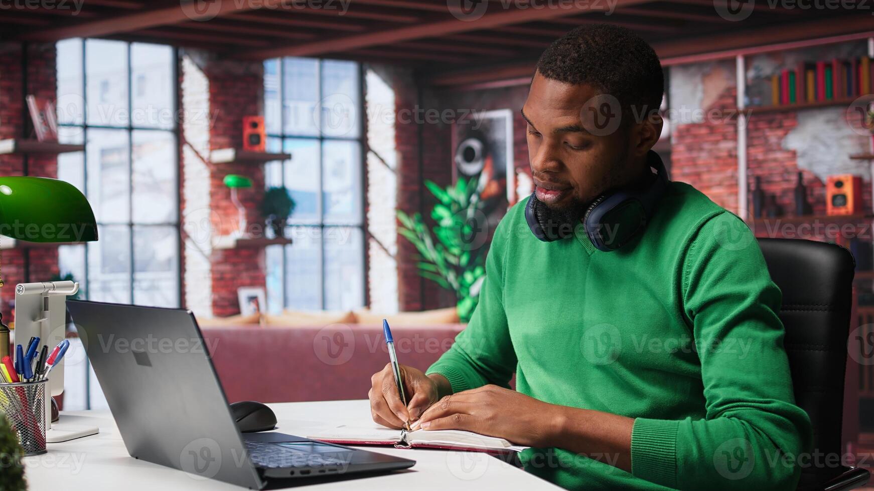 African american teleworker writing personal notes at desk in modern loft, remote job. Productive guy sitting at home office, using pen to take notes in textbook. Camera B. photo