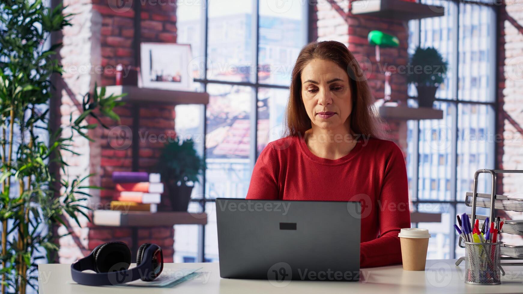 Mature girl freelancer managing her creative projects remotely in loft, working on a new novel or script for her gig. Woman focusing on writing and editing her creation, telework. Camera A. photo