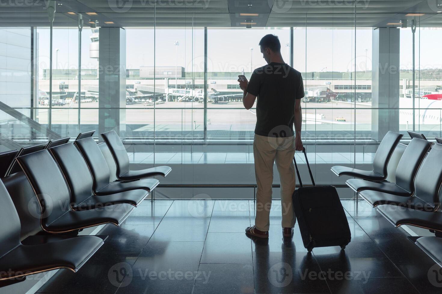A man stands at a large airport window, holding his suitcase and checking his phone while waiting for his flight. Concept of solitude, anticipation and modern travel technology. photo