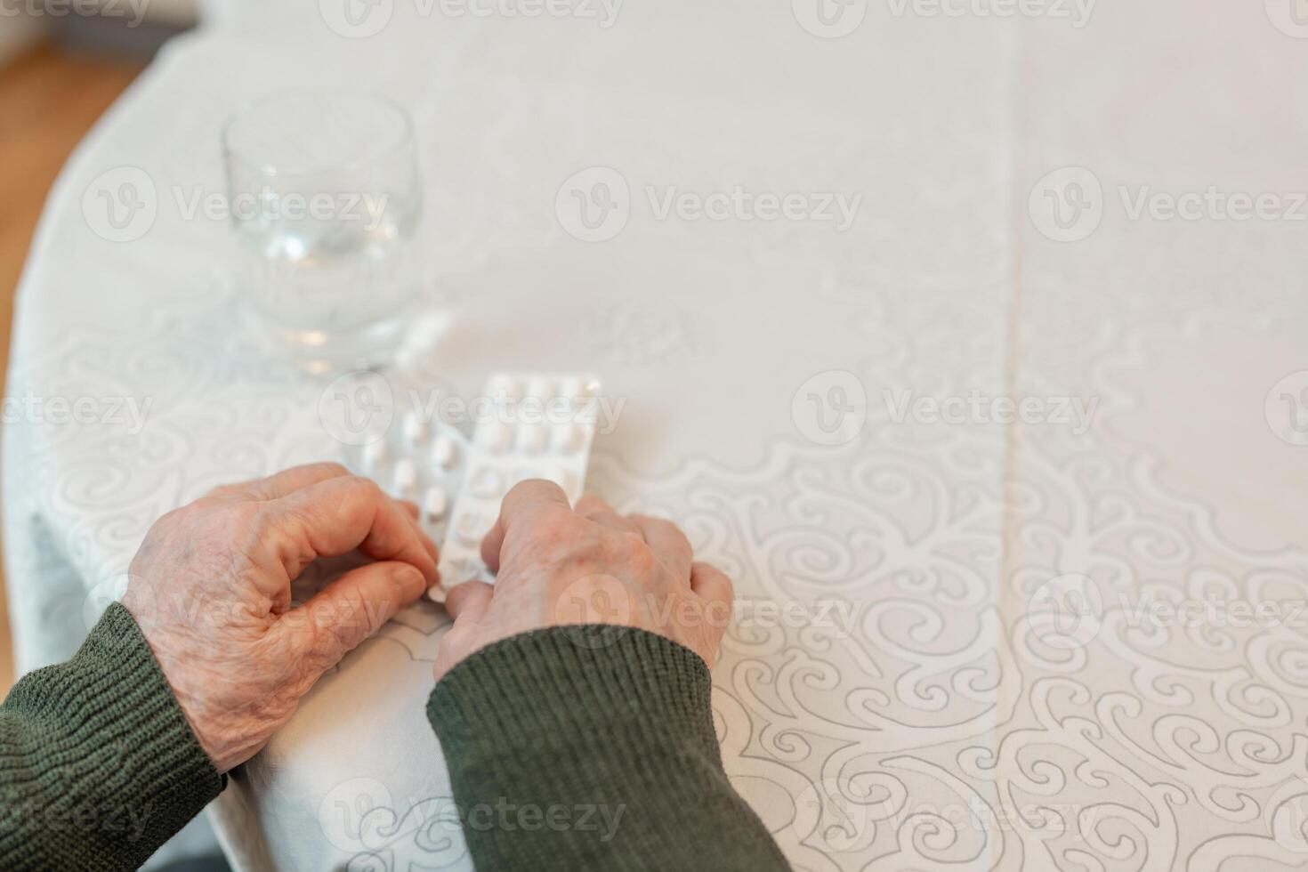An elderly person hands holding blister packs of pills over a patterned tablecloth, with a glass of water nearby, copy space. Concept of senior health care and daily medication routine photo