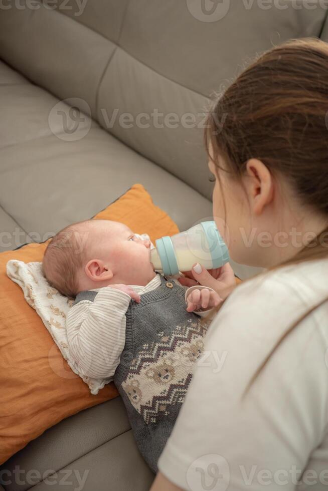 A young mother is bottle-feeding her baby on a cushion on a sofa at home. Concept of infant care, nurturing, early motherhood, feeding routine, and emotional connection in a home setting. photo