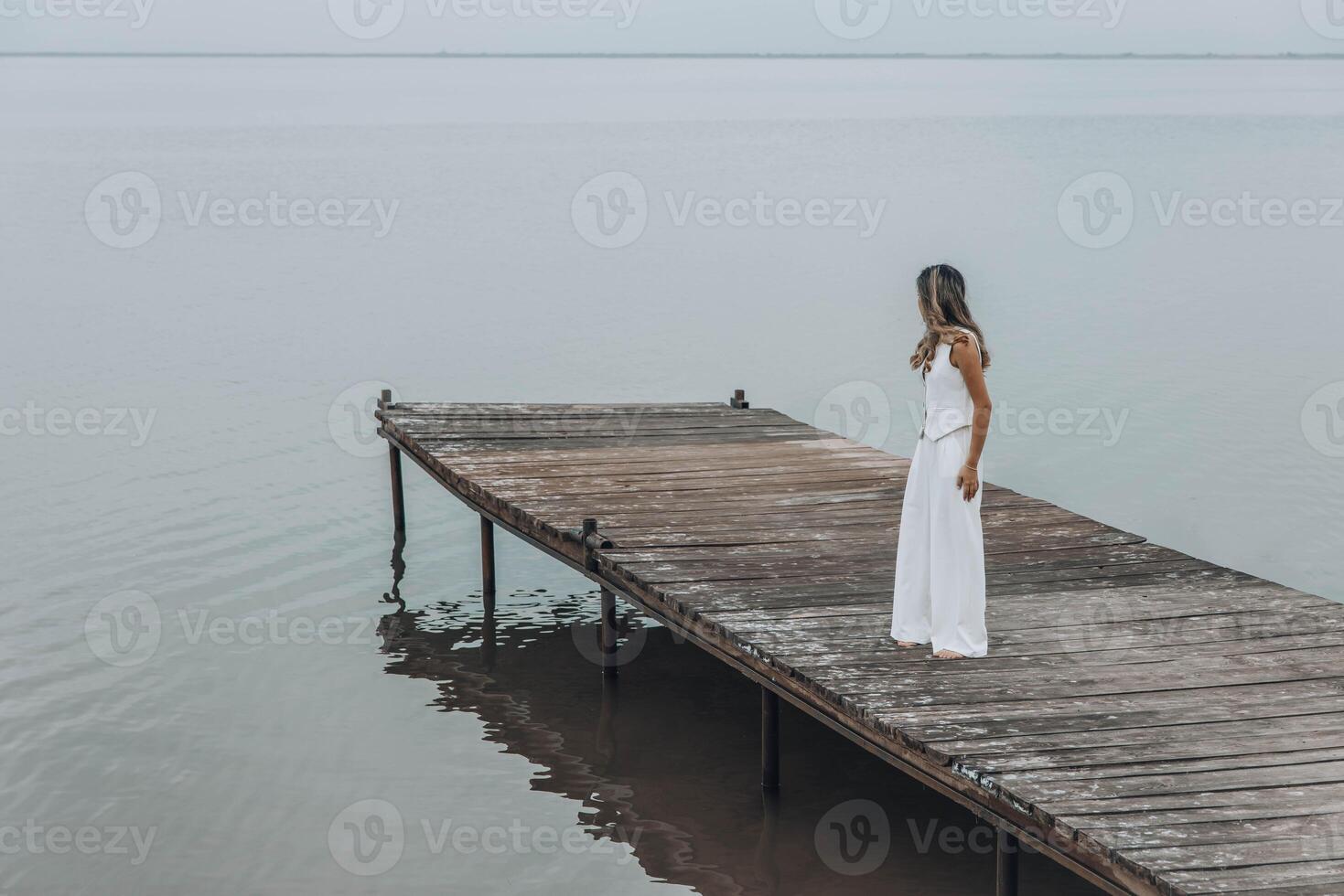 unrecognizable woman in white outfit stands on wooden pier, calm lake, peaceful overcast, viewed from behind with contemplative mood, mindfulness, travel retreats, wellness photo