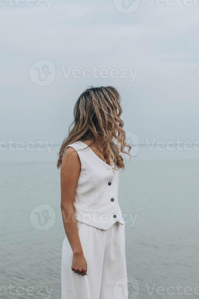unrecognizable woman with wavy hair in a white outfit stands by a still lake, gazing to the side. the image conveys a serene and contemplative mood, emphasizing inner peace and self connection. photo