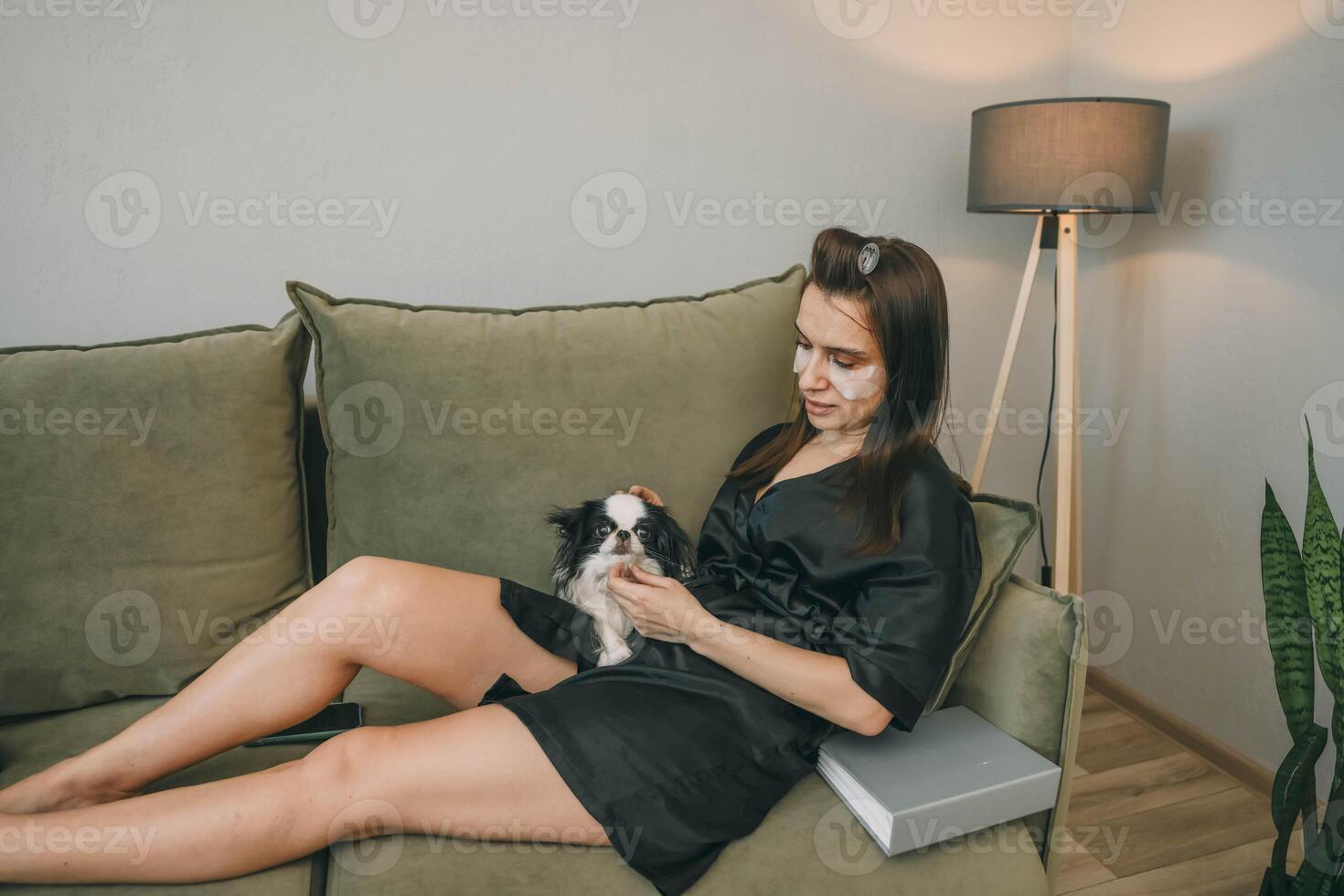 woman with eye patches relaxing on couch with book, and petting dog in cozy living room, concept of self-care and pet care photo