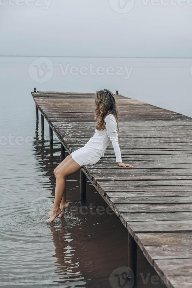 unrecognizable woman in a white dress, sitting on the edge of wooden pier, dipping her feet into calm water with a light splash, gazing at horizon under soft cloudy sky, relaxation, travel, vertical photo