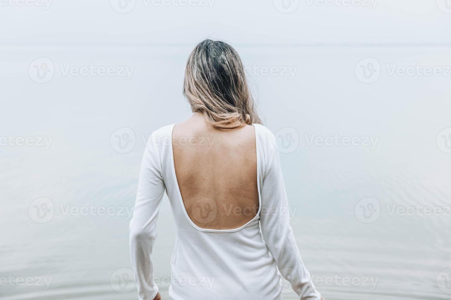 unrecognizable woman standing with back exposed in open white dress near calm lake, facing the water with relaxed posture and serene mood, concept of wellness, spa, fashion editorial photo