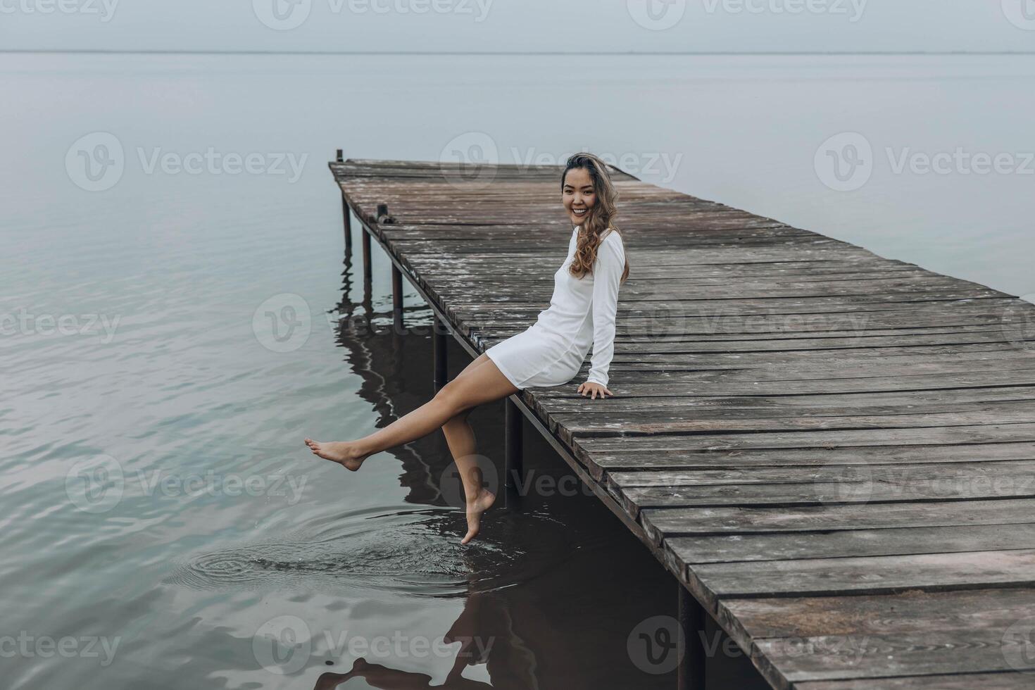 Asian woman in a white dress, sitting on the edge of wooden pier, dipping her feet into calm water with a light splash, gazing at horizon under soft cloudy sky, relaxation, travel photo