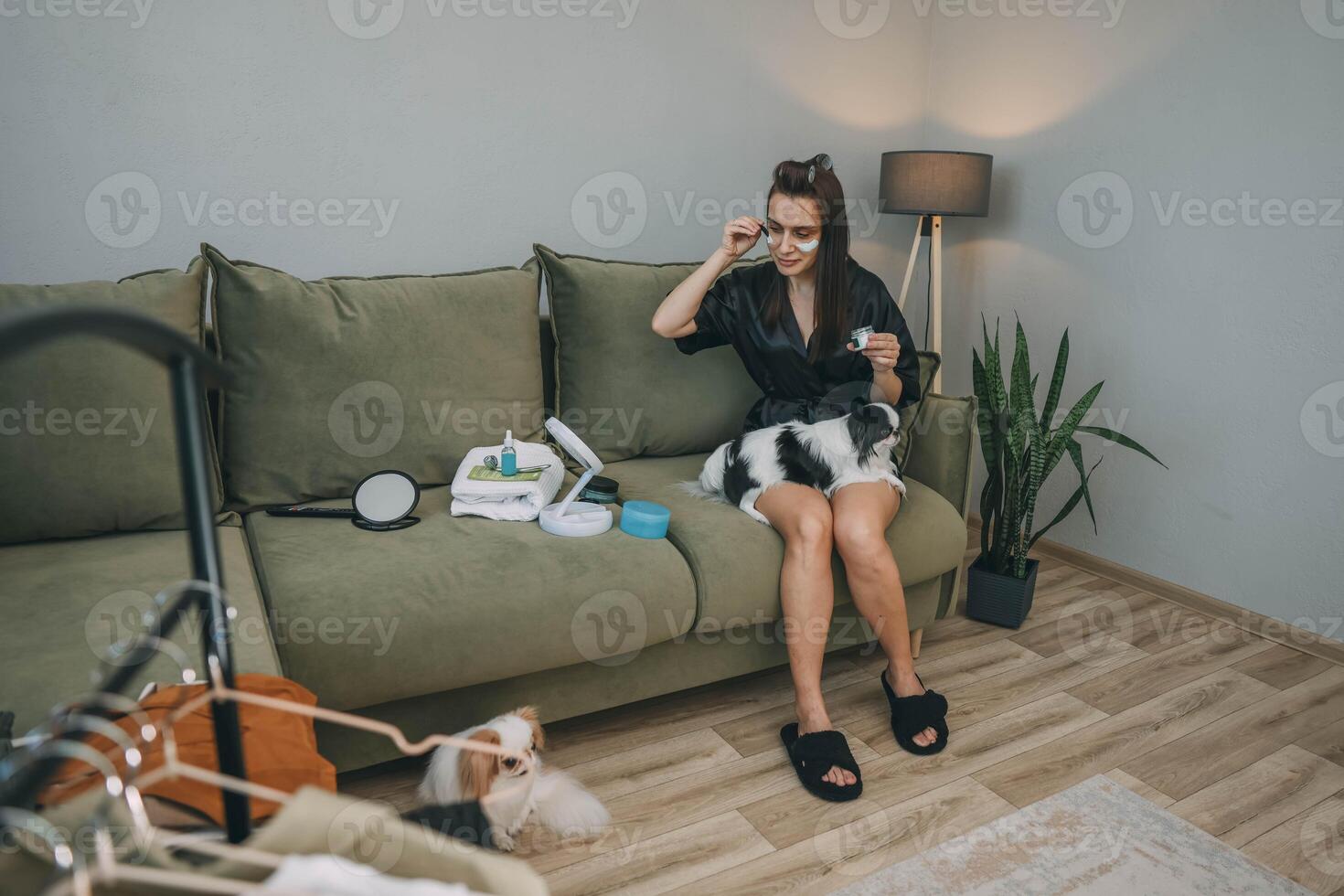 woman applying face cream under eyes with one japanese chin dog on lap, another sits on floor in cozy living room with plant, wellness lifestyle, pet care, beauty product photo