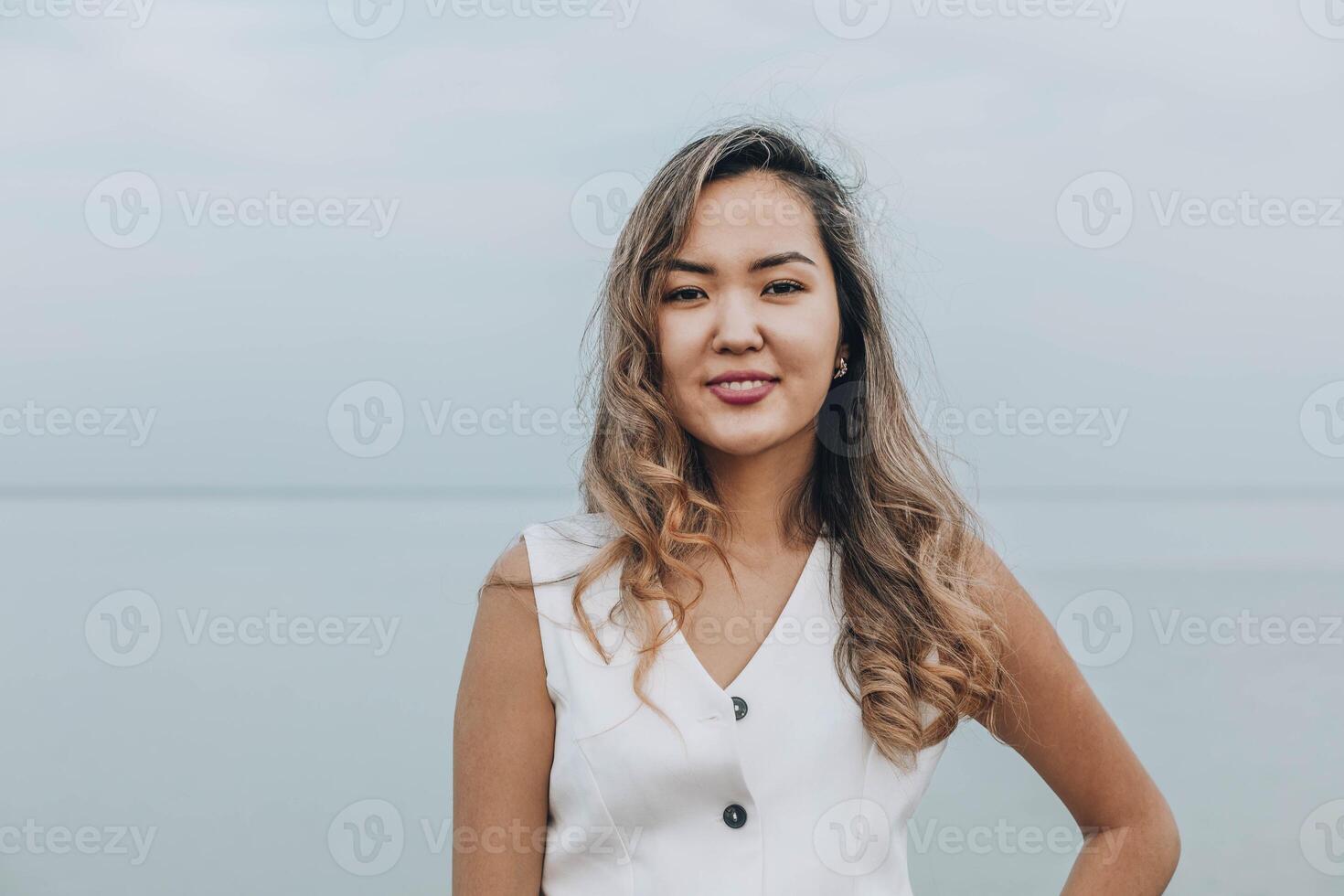 Asian woman smiling and looking at camera on a calm lakeside, dressed in white outfit, displaying a confident and cheerful emotion, concept of wellness, skincare, personal development photo