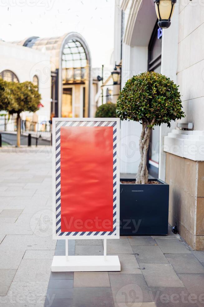 Blank advertising board stands on sidewalk near trees and modern architecture, daytime photo