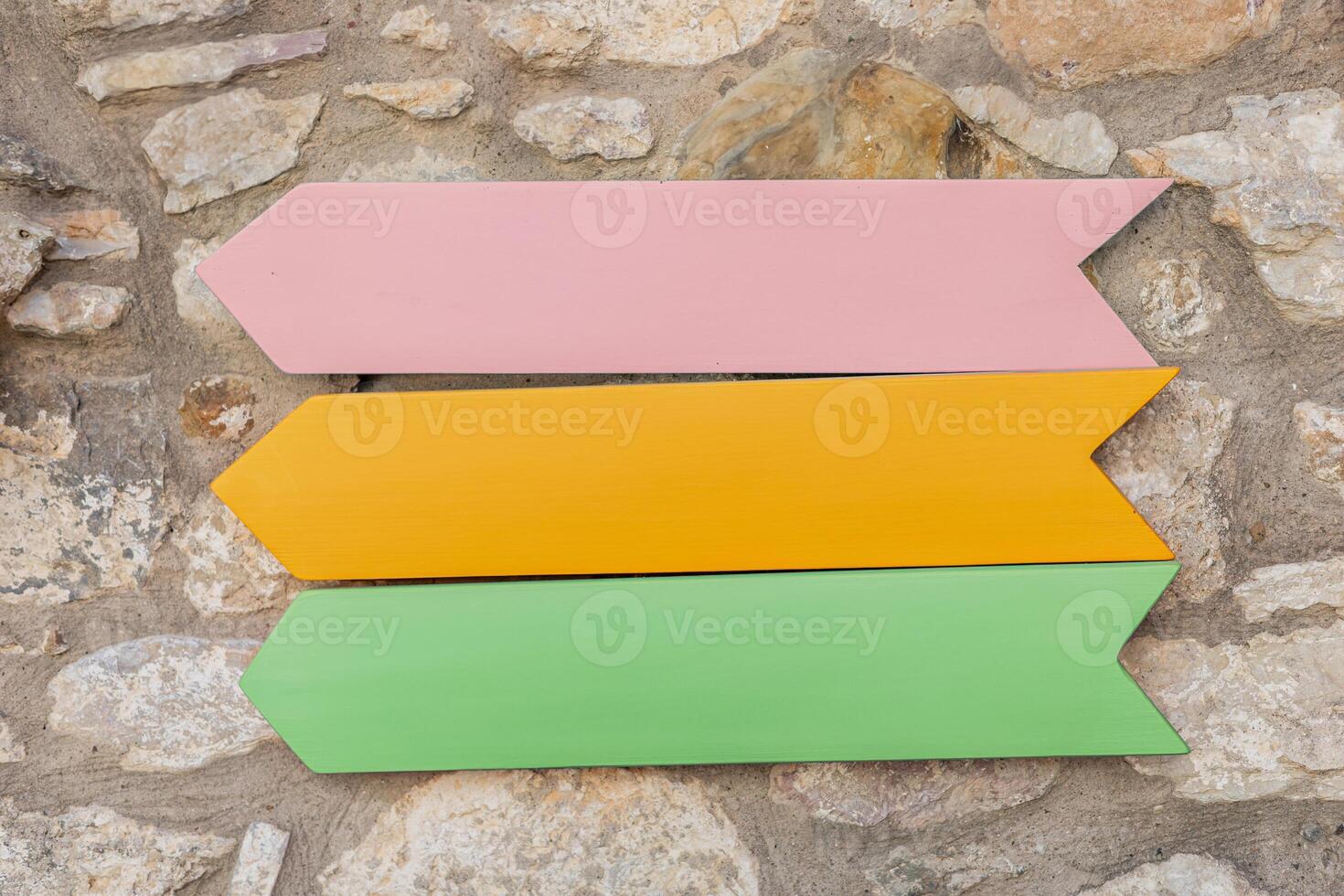 Vibrant wooden mockup arrows in pink, yellow, and green are displayed against a textured stone wall outdoors under natural light. photo