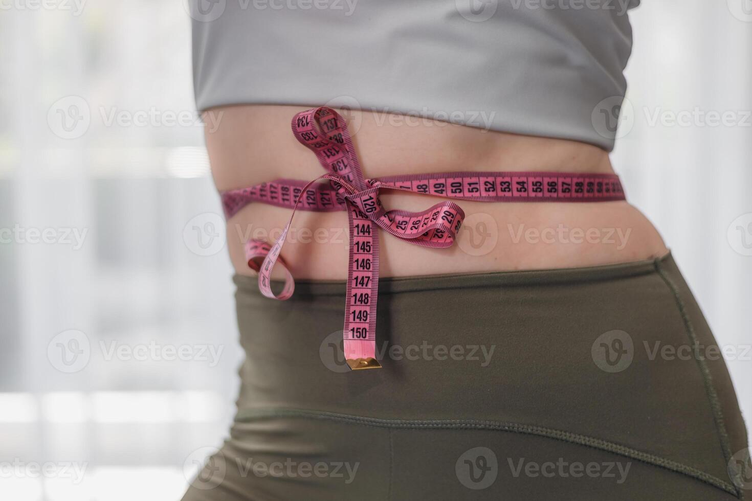 A close-up of a womans waist with a measuring tape tied around it. The woman has a slim figure and is wearing top and leggings. photo