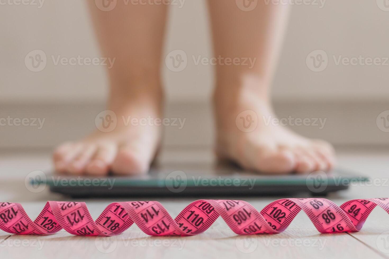 A pair of feet stands on a digital scale. A pink measuring tape is coiled in front of the scale. focusing on health and fitness. photo