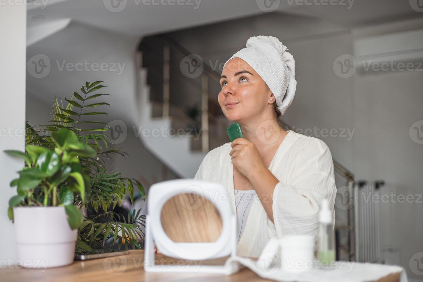 young Caucasian woman with a towel performs gua sha face massage in a bright room. She holds a jade tool and looks relaxed. photo