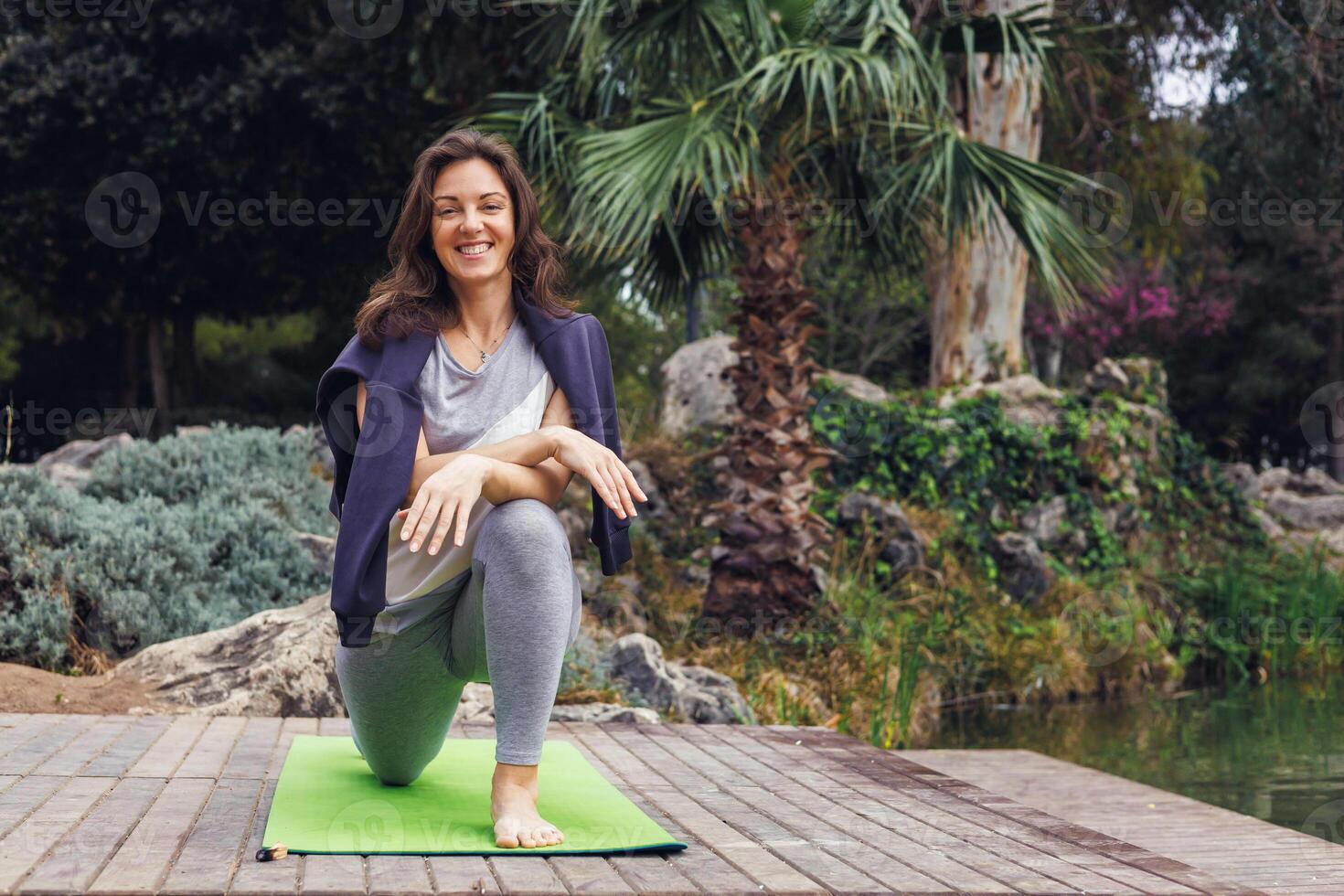 Caucasian woman with long brown hair practices yoga on green mat in a park. Palm trees and shrubs are visible in the background. photo