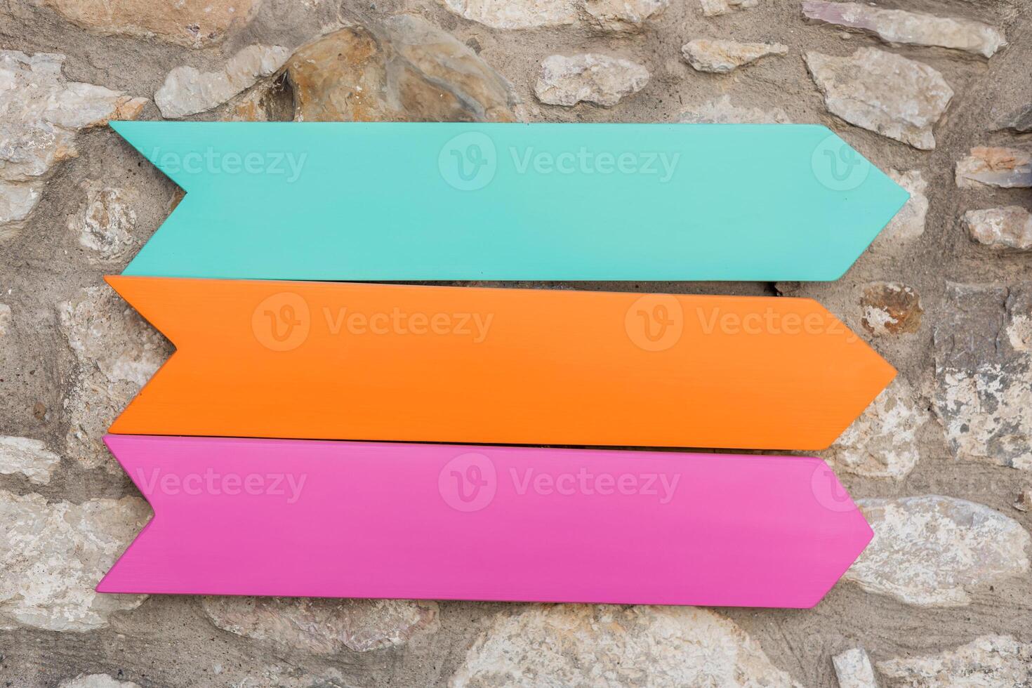 Vibrant wooden mockup arrows in pink, orange, and blue are displayed against a textured stone wall outdoors under natural light. photo