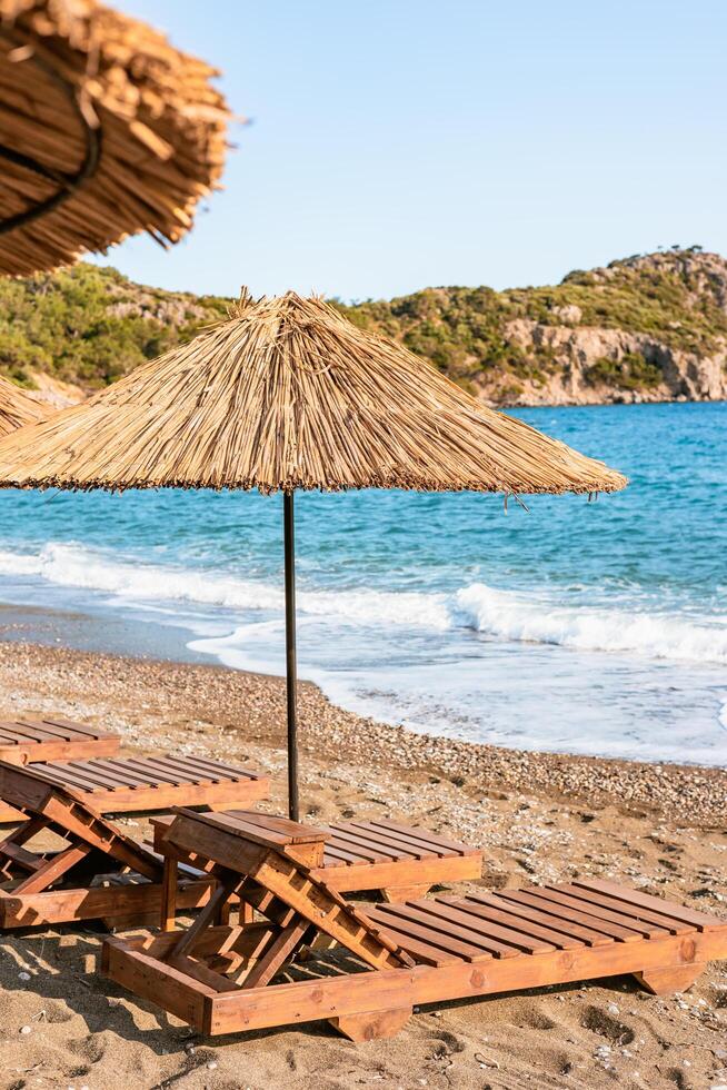 Wooden loungers with straw umbrellas line the sandy beach while gentle waves lap at the shore under a clear blue sky. photo