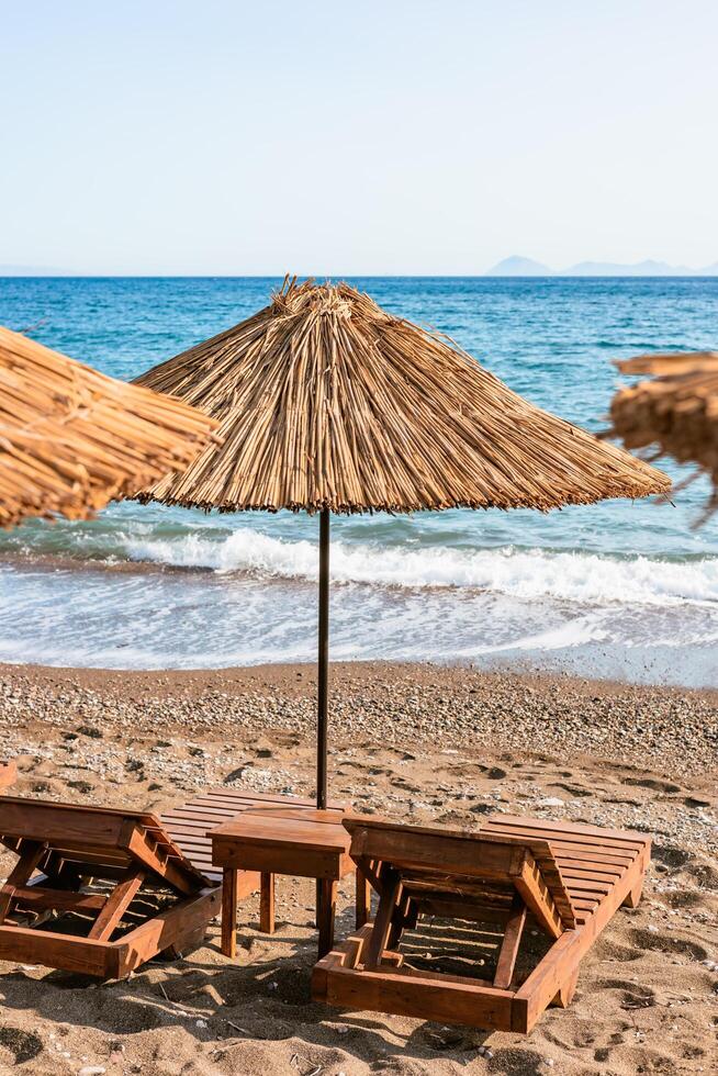 Wooden lounger with straw umbrellas line the sandy beach while gentle waves lap at the shore under a clear blue sky photo