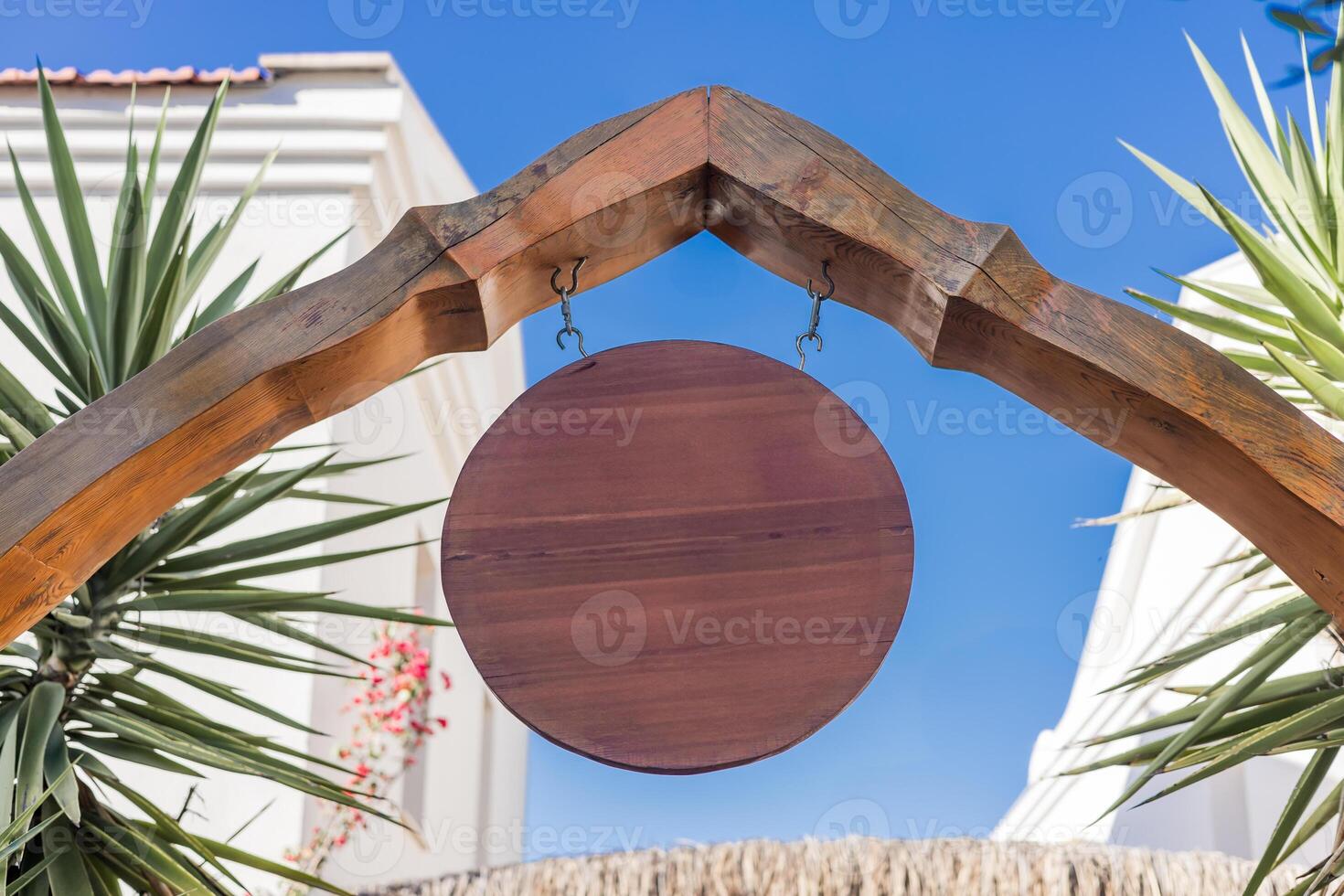 Wooden sign hanging from arch in sunny outdoor setting surrounded by greenery and buildings. Mockup for shop, cafe or restaurant photo