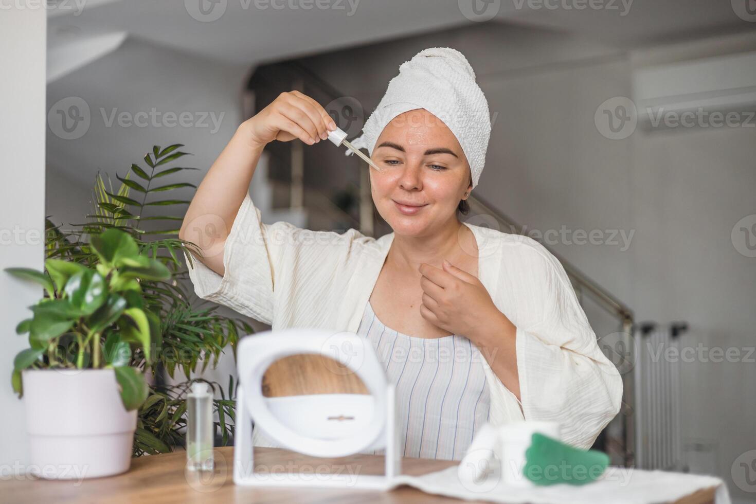 A young Caucasian woman with brown hair in a towel applies serum to her face while preparing for a gua sha massage. photo
