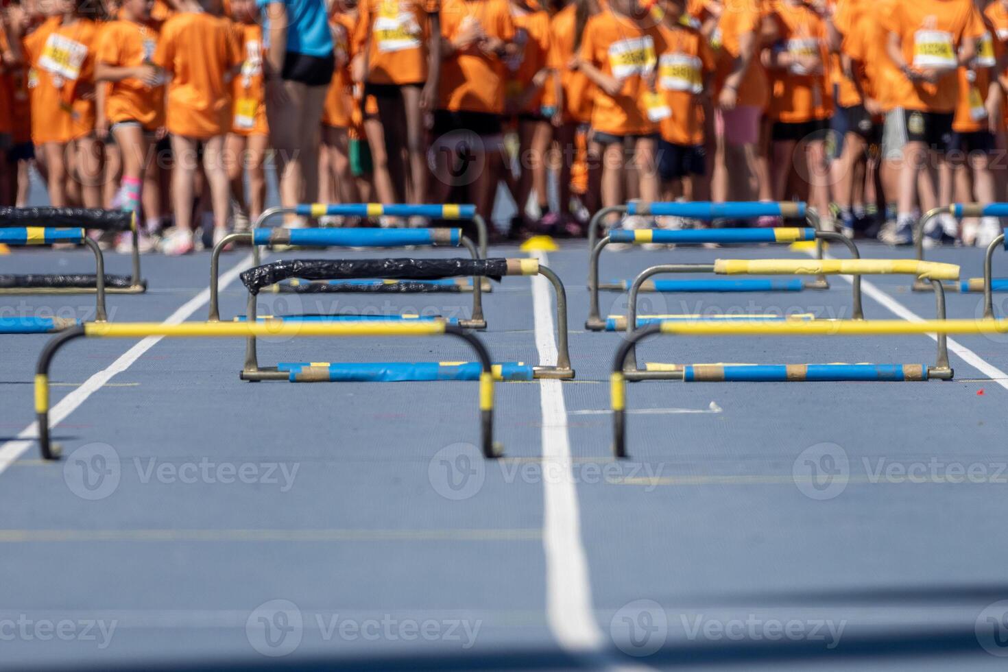 Blue athletics track with yellow and black hurdles aligned and young athletes waiting in the background photo