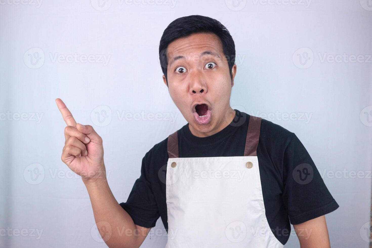 Funny shocked facial expression young asian man wearing apron pointing right side, empty space. photo