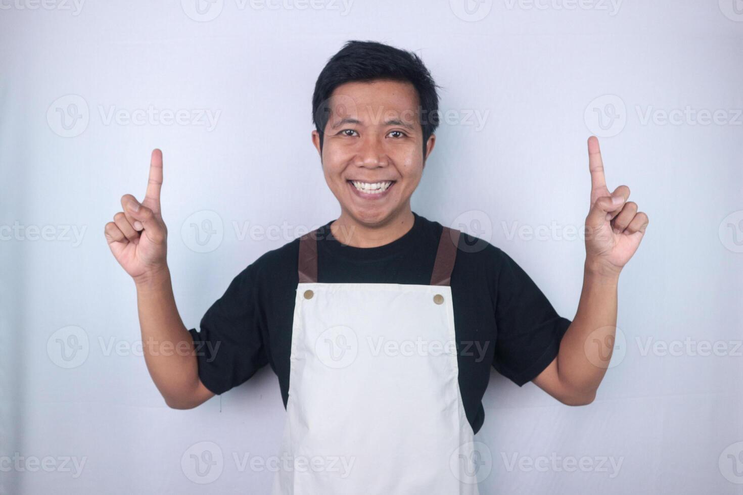Smiling face expression young asian man wearing apron pointing up, empty space, isolated white background photo