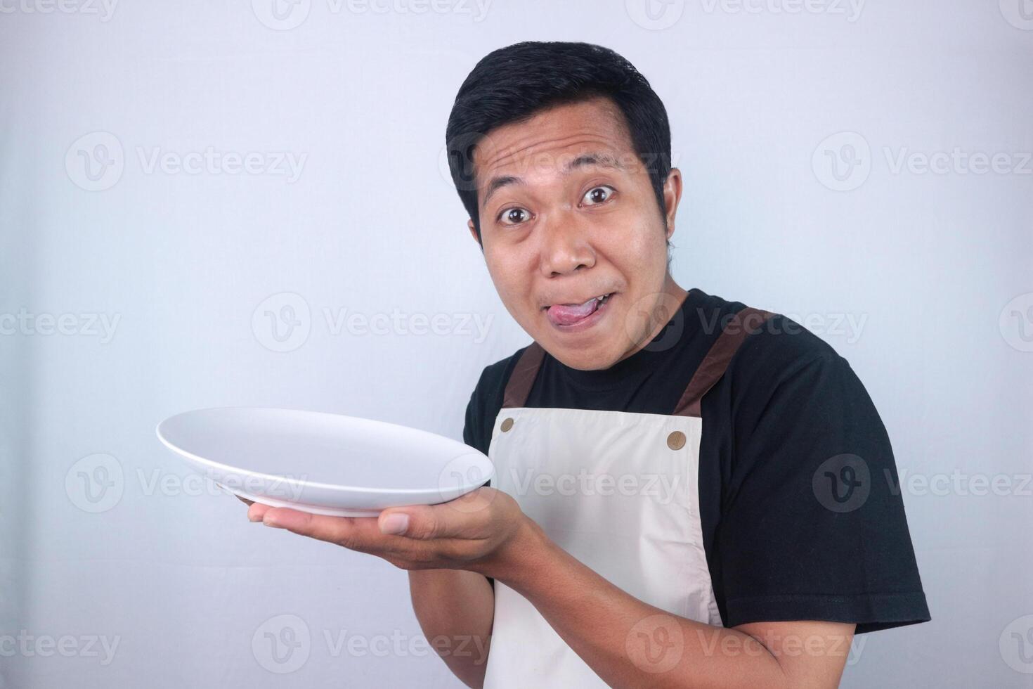 Funny expression asian young man showing delicious dish on white plate while stuck out his tongue. photo