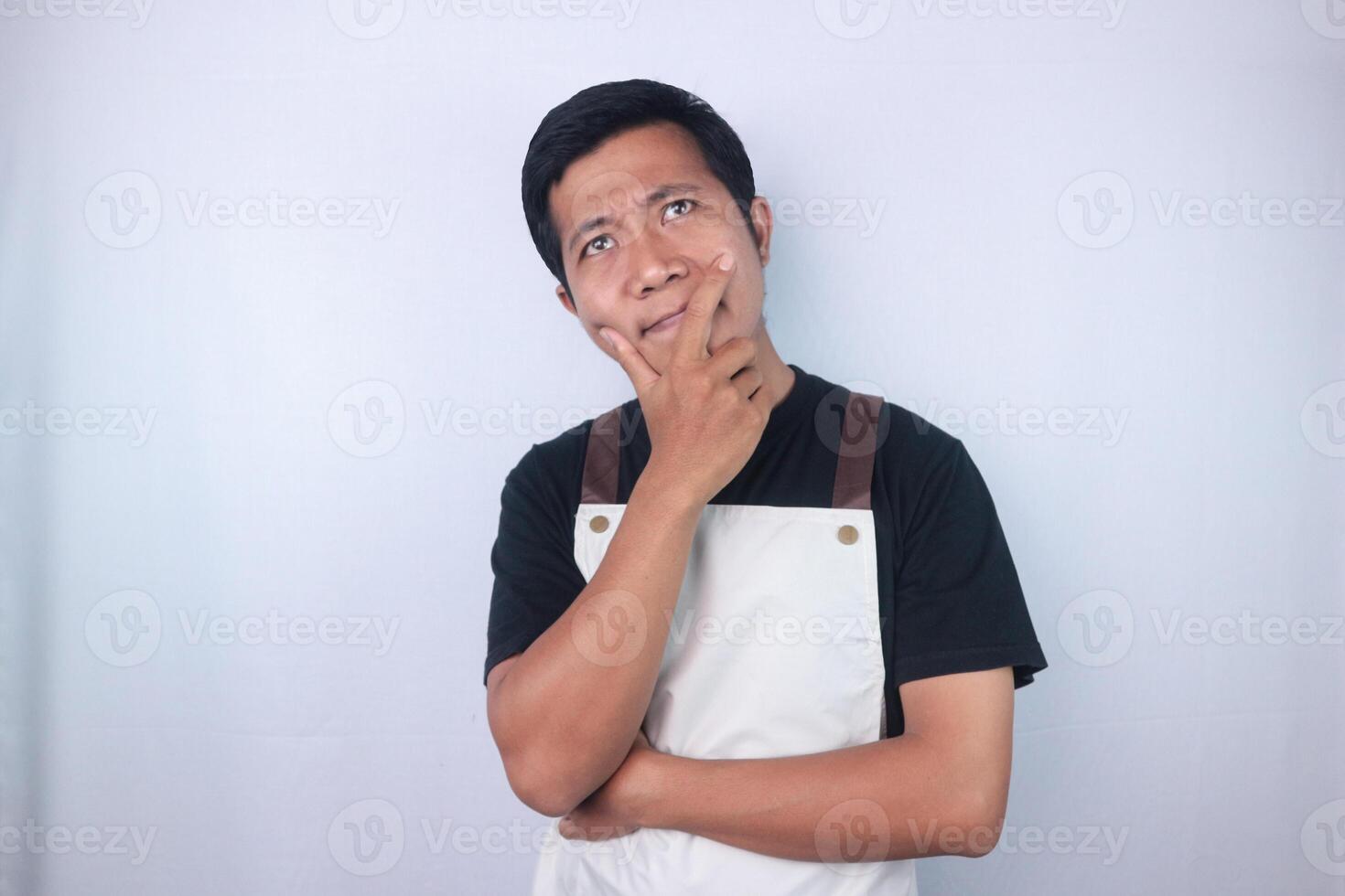 Young asian man wearing apron gesture thinking about something looking at copy space photo