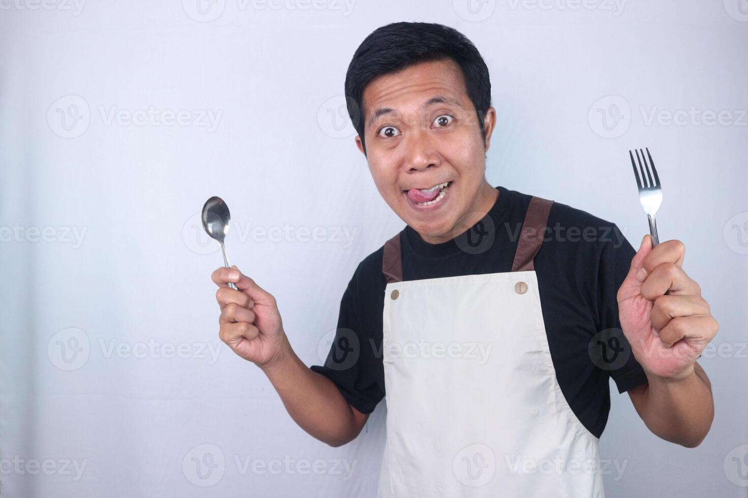 Funny expression asian young man hold spoon and fork while stuck out his tongue, ready eating dish. photo