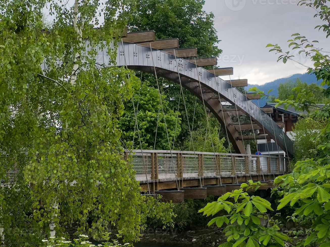 A modern footbridge spans a tranquil river, set against a backdrop of verdant trees and mountains. photo