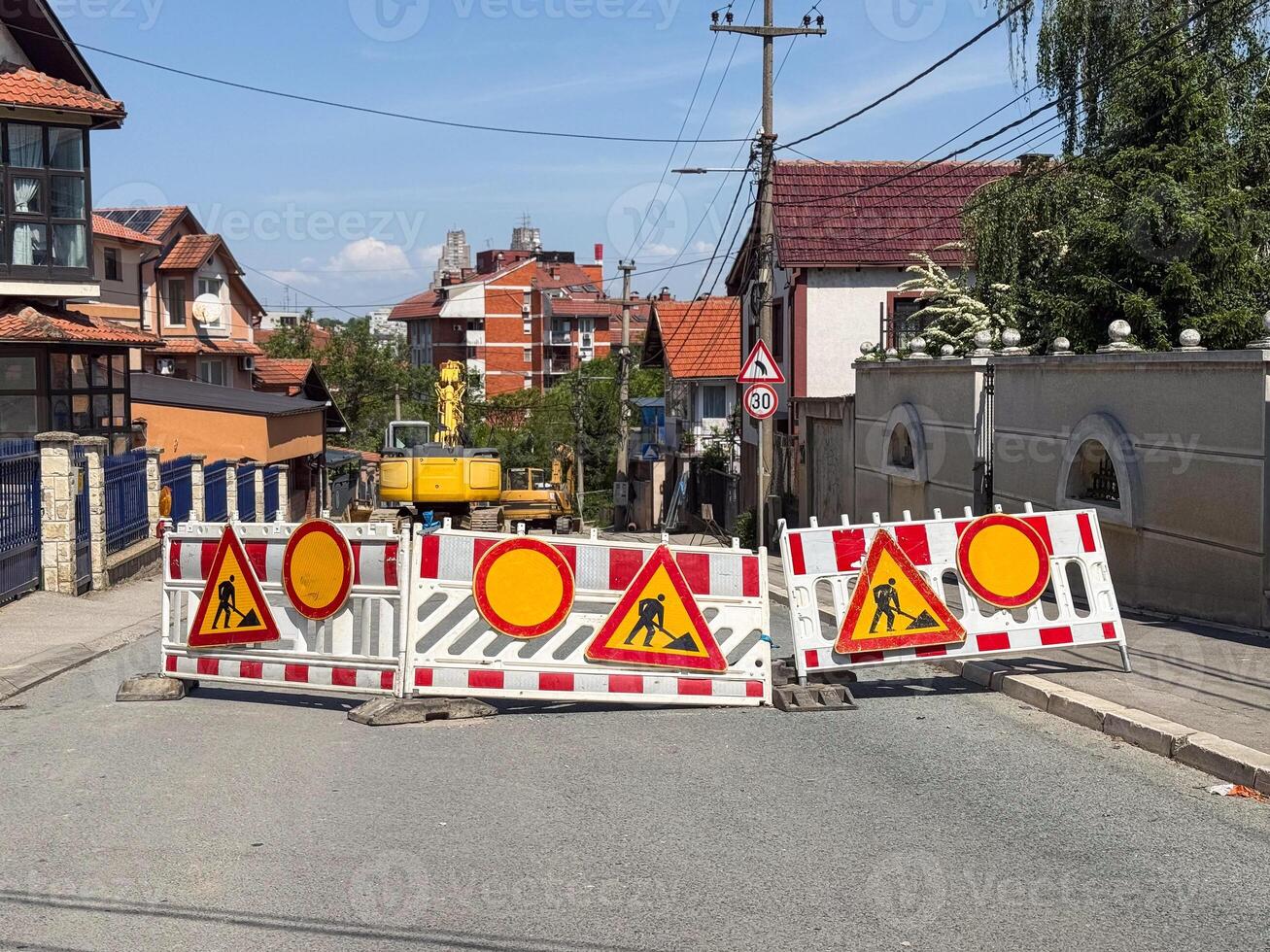 Street closed for construction work. Traffic barriers with warning signs and excavator visible in residential area. photo