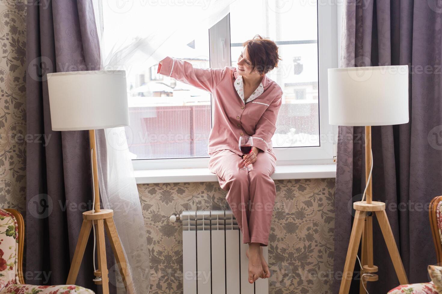 Beautiful woman wears pajamas with a glass of red wine sits on the windowsill near the beautiful window at home. photo