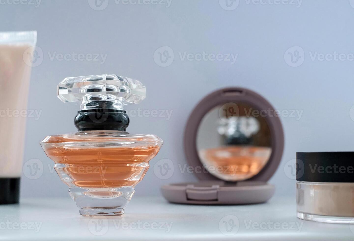 Elegant flat lay of a perfume bottle with peach-colored fragrance, compact mirror reflecting the bottle, and makeup items on a white surface. photo