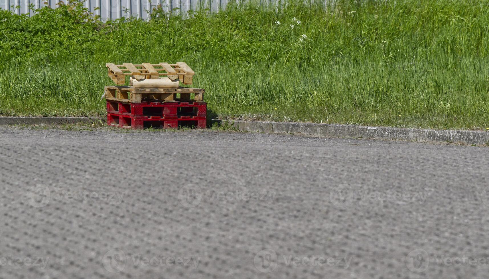 Red and pure wooden pallets are stacked on a driveway in front of a green area, the foreground and paved photo